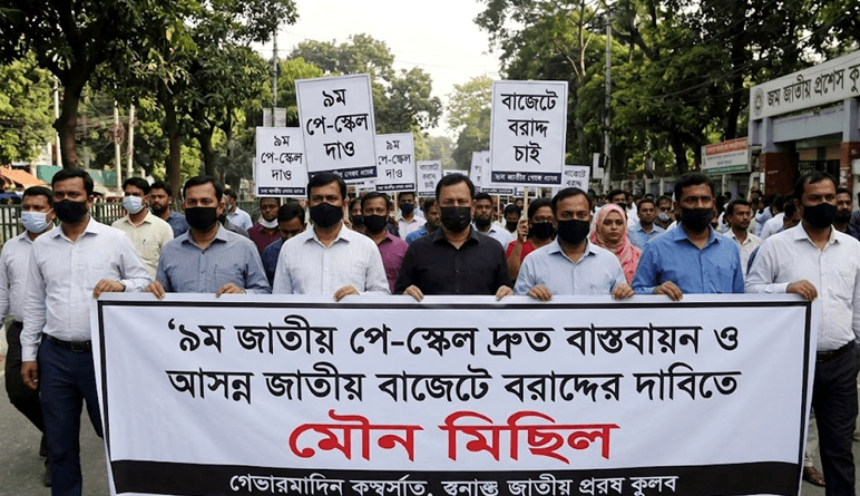 Crowd of Protesters Wearing Masks March Down a Street Holding a Large Bengali Banner