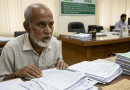 Older Man with Gray Hair and Beard Sits at a Desk in an Office Reviewing a Large Stack of Papers with a Focused Expression