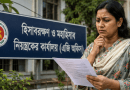 Woman in a floral shawl studies a paper outside a blue Bengali government office sign.