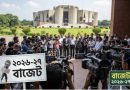Group of reporters and camera crews surround officials for a budget briefing outside a government building. Bengali signs are visible in the foreground and a large monument is in the background.