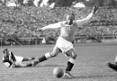 Soccer player in mid-kick on a grassy field, ball near his foot, another player sliding on the ground nearby in a vintage match scene.