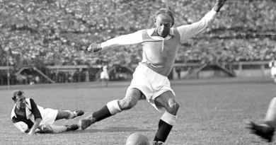 Soccer player in mid-kick on a grassy field, ball near his foot, another player sliding on the ground nearby in a vintage match scene.