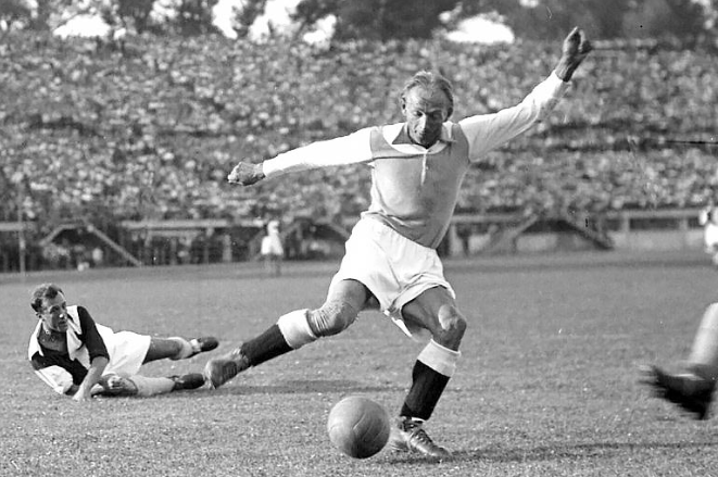 Soccer player in mid-kick on a grassy field, ball near his foot, another player sliding on the ground nearby in a vintage match scene.